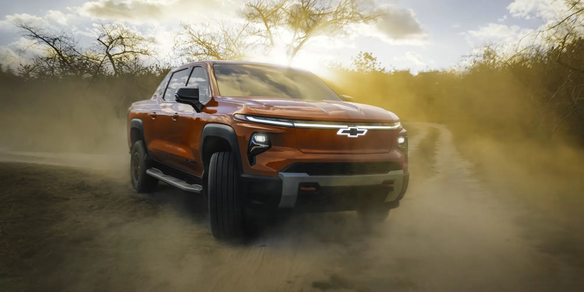 Chevrolet Silverado EV 2026 orange foncé roulant sur un chemin poussiéreux, phares allumés, symbolisant ses performances en terrain difficile.