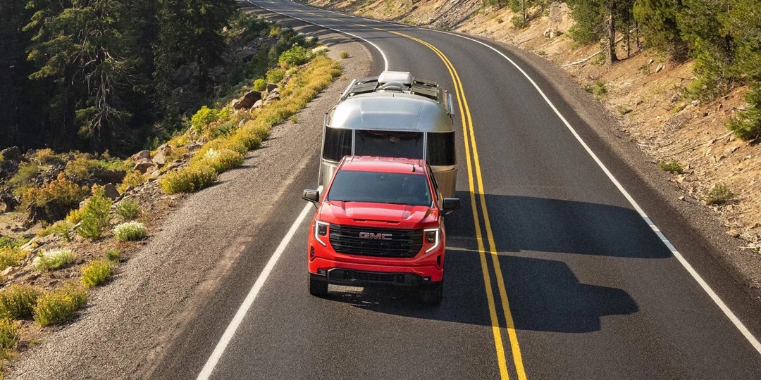 GMC Sierra 1500 2026 rouge tractant une roulotte argentée sur une route de montagne, illustrant sa puissance de remorquage.
